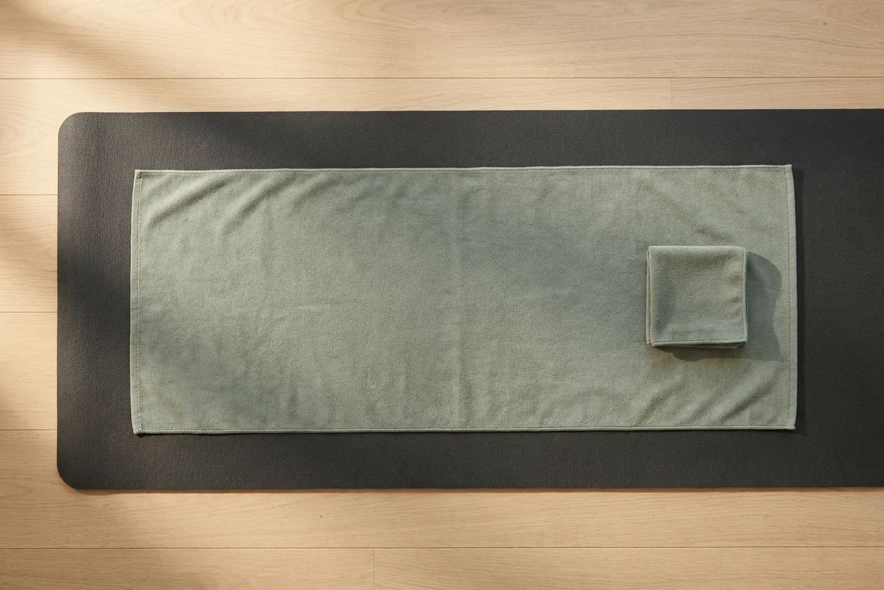 A mat-length microfibre yoga towel laid beside a small folded hand towel on a yoga mat, showing the size difference