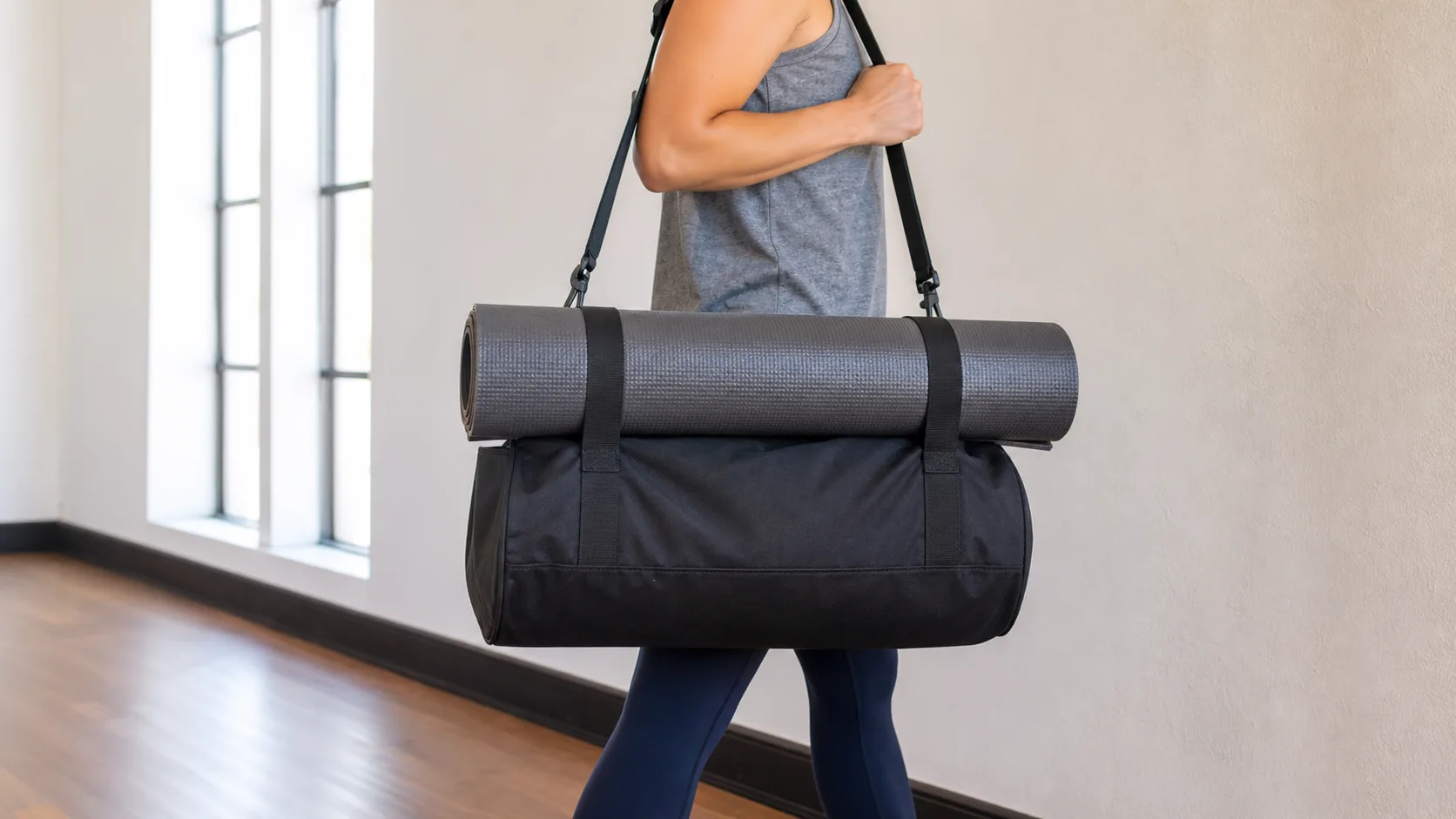 Person carrying a black gym duffel with a centered rolled yoga mat held horizontally by two spaced straps