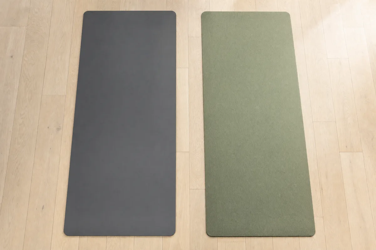 Two premium yoga mats side by side on a wooden studio floor — a dense dark charcoal mat and a sage-green rubber mat