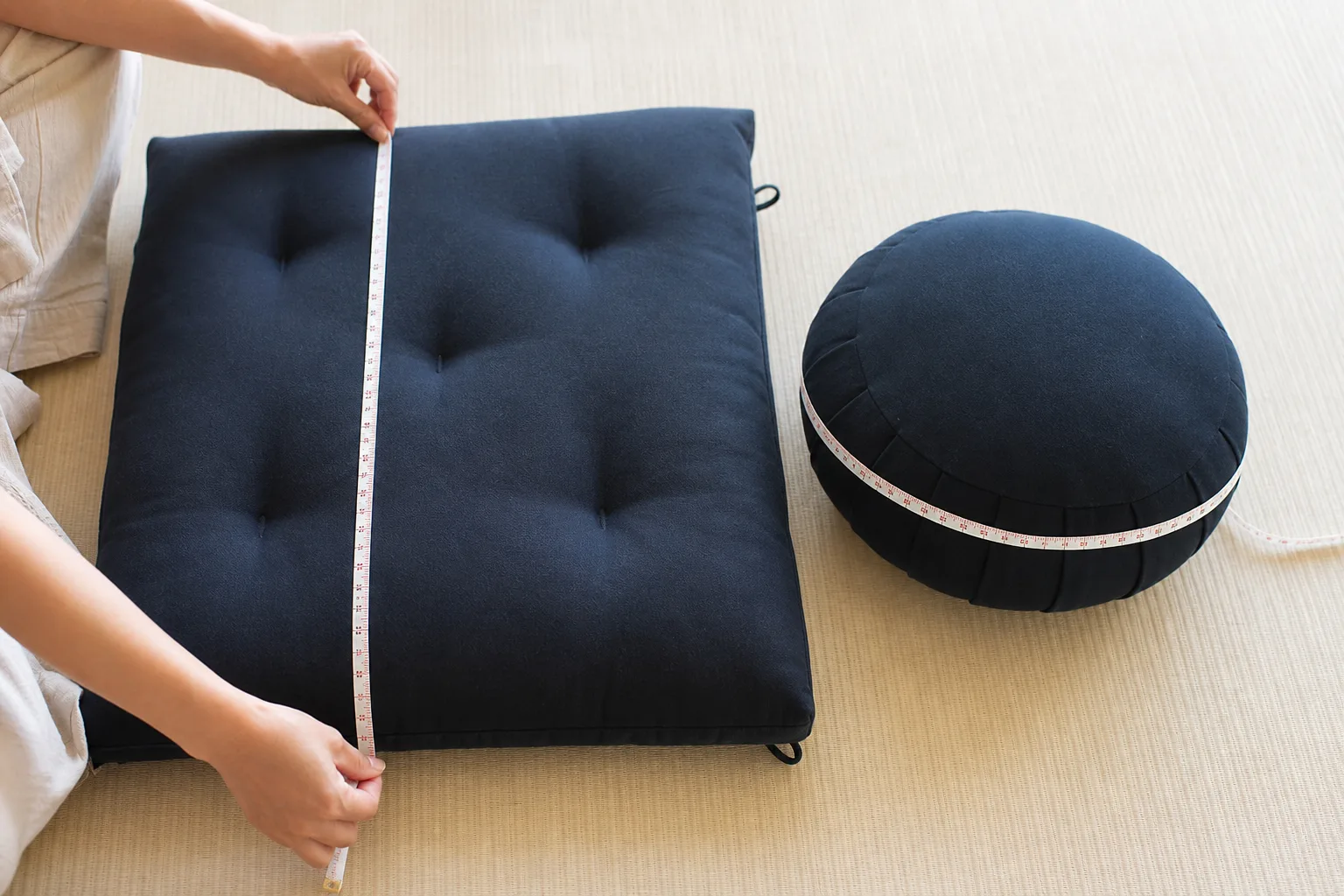 Realistic hands measuring a dark rectangular zabuton length and matching round zafu diameter with a flexible tape