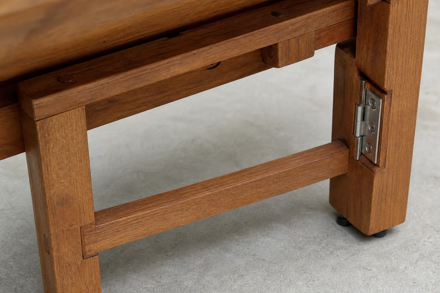 Close underside view of a low wooden table showing leg joinery, cross brace, hinge, fasteners and anti-slip feet