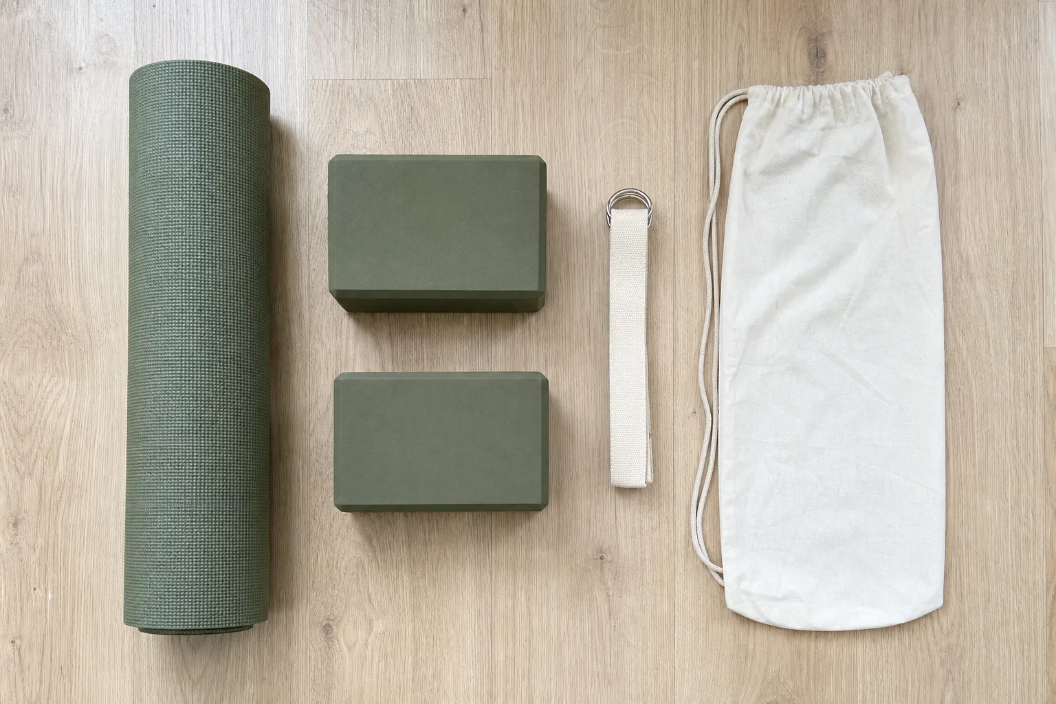 Overhead editorial photograph of one rolled green yoga mat, two matching green blocks, one cream D-ring strap and one empty drawstring bag spaced separately
